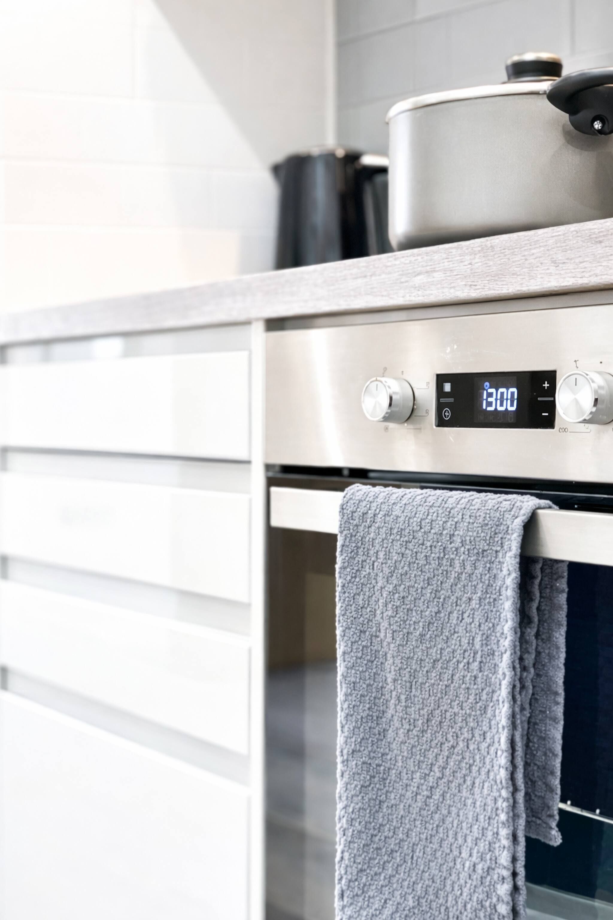 A modern kitchen with stainless steel appliances, a digital clock displaying 13:00, and a towel hanging on the oven handle.
