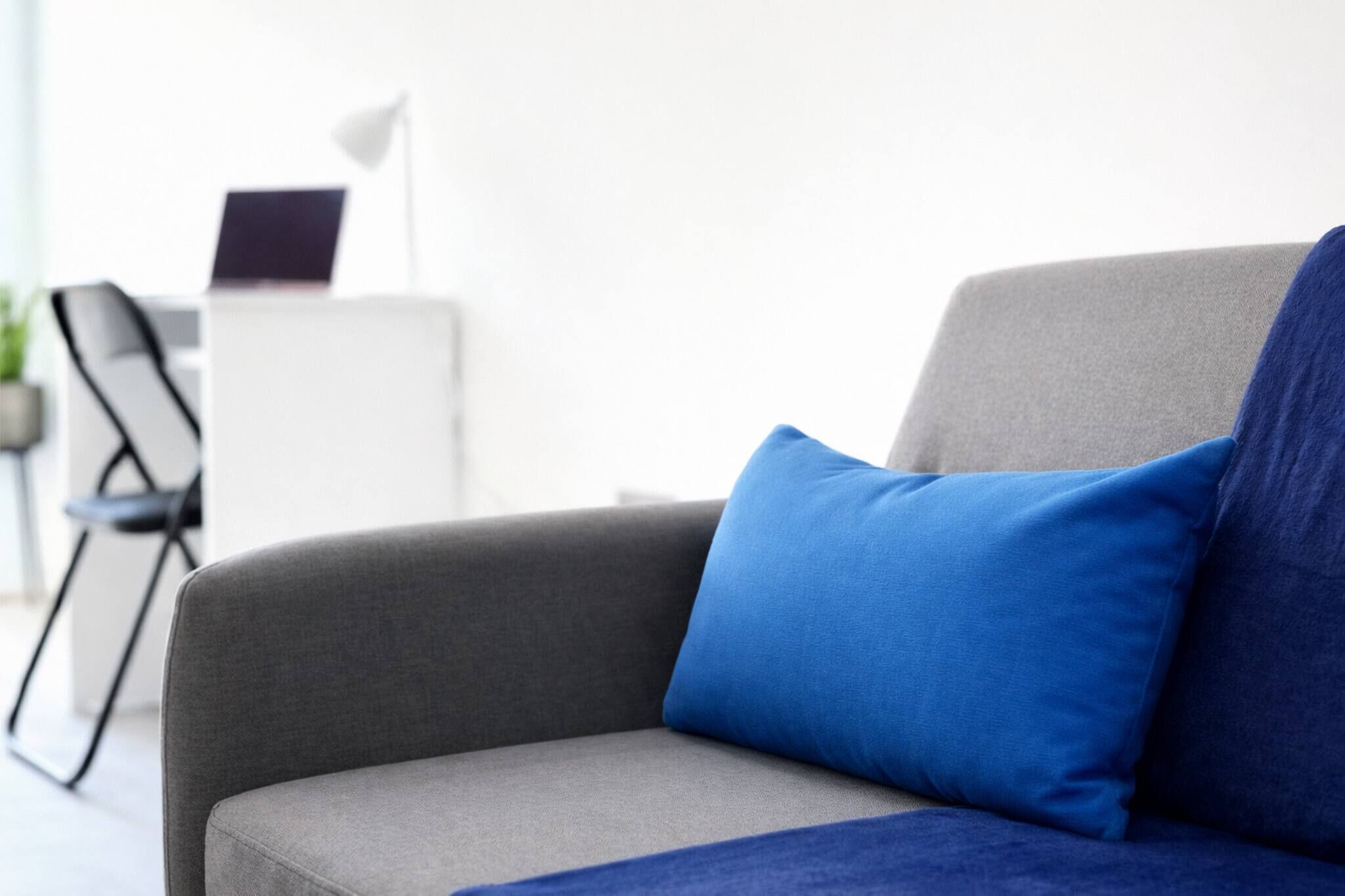 Ein graues Sofa mit blauen Kissen in einem modernen Raum, der einen weißen Schreibtisch und einen Laptop enthält.