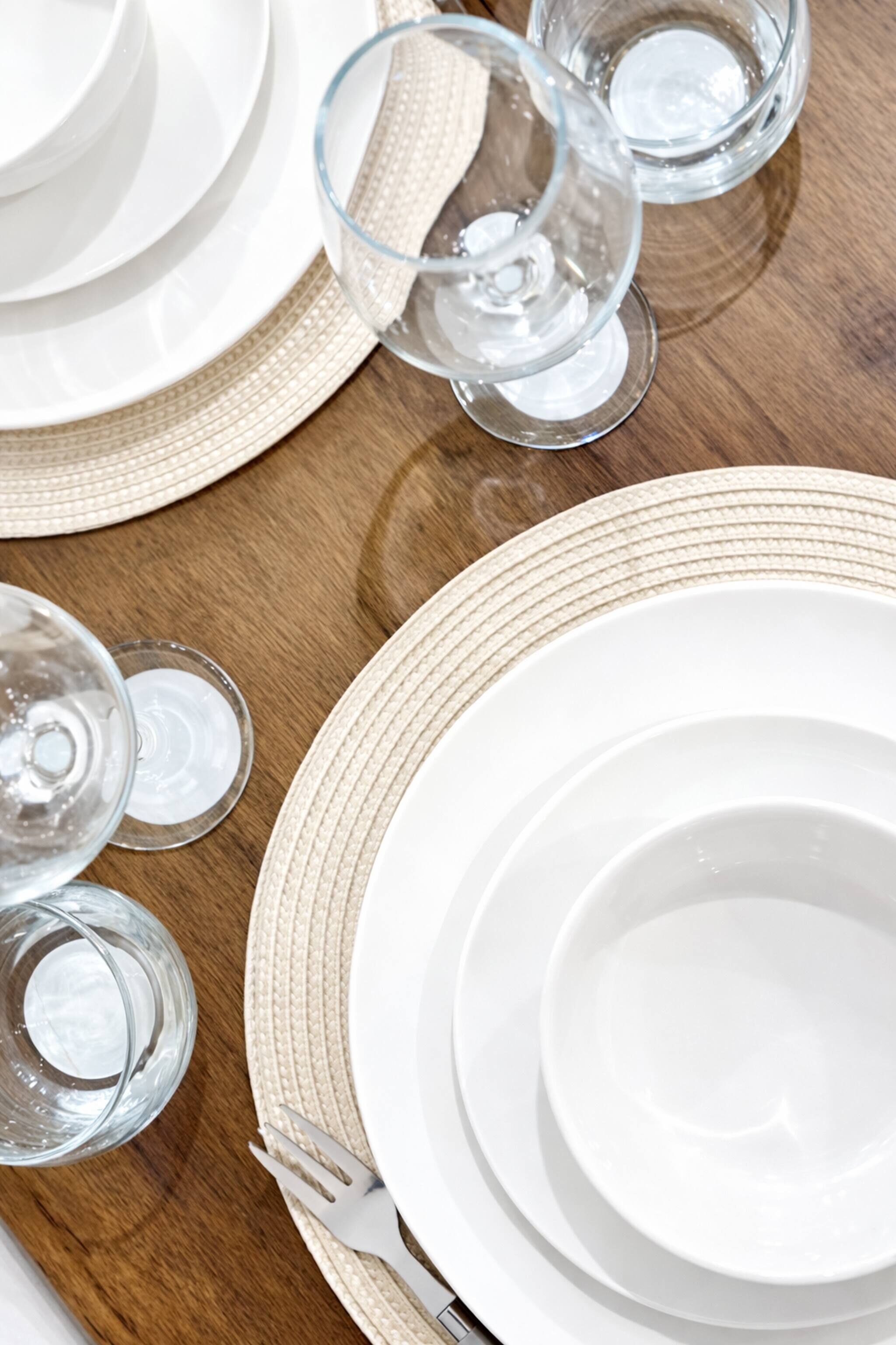 A dining table set with white plates, clear glasses, and a woven placemat.