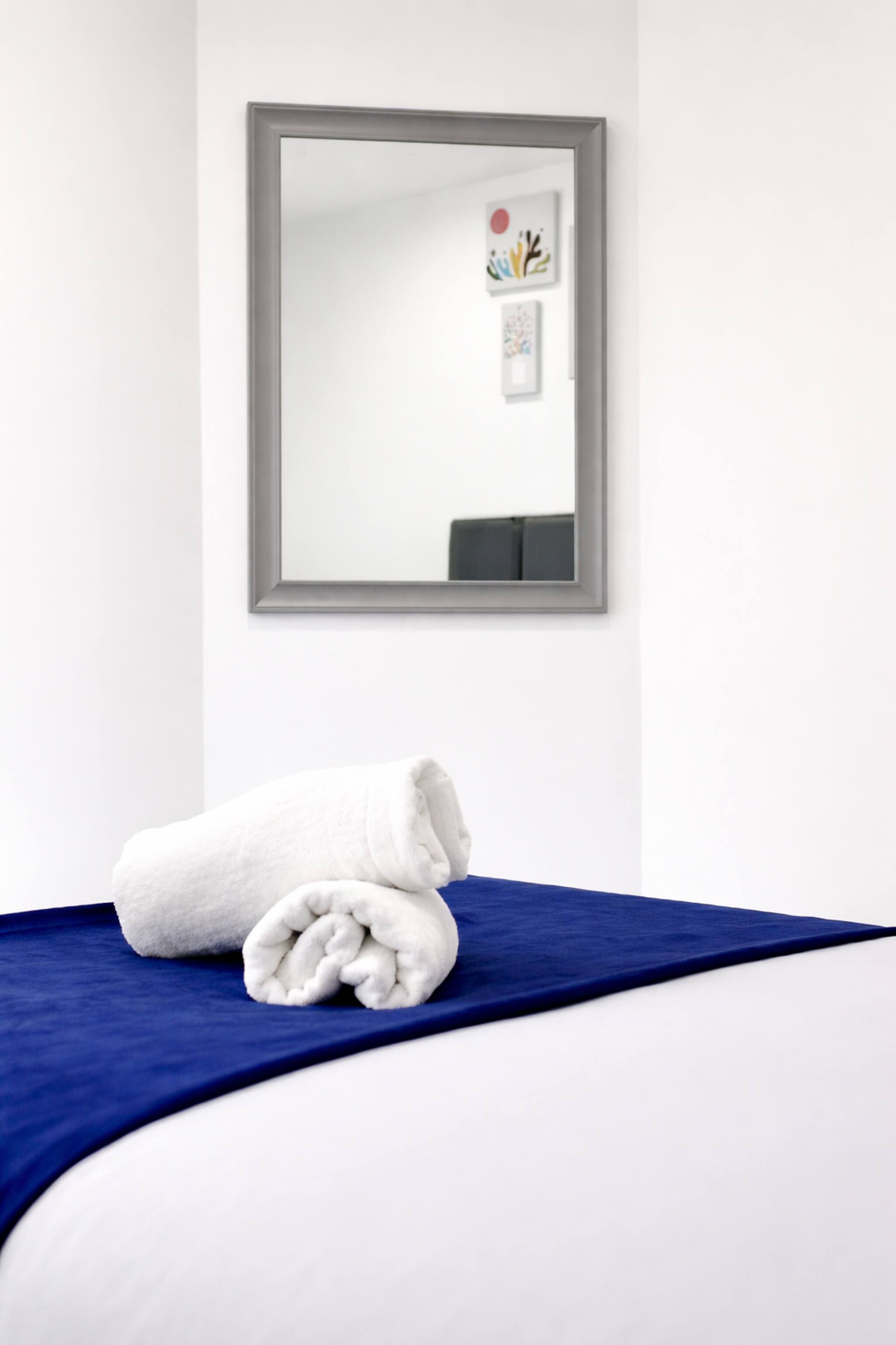 A neatly made bed with a white comforter and a blue blanket, a white towel folded on the bed, and a framed mirror on the wall.