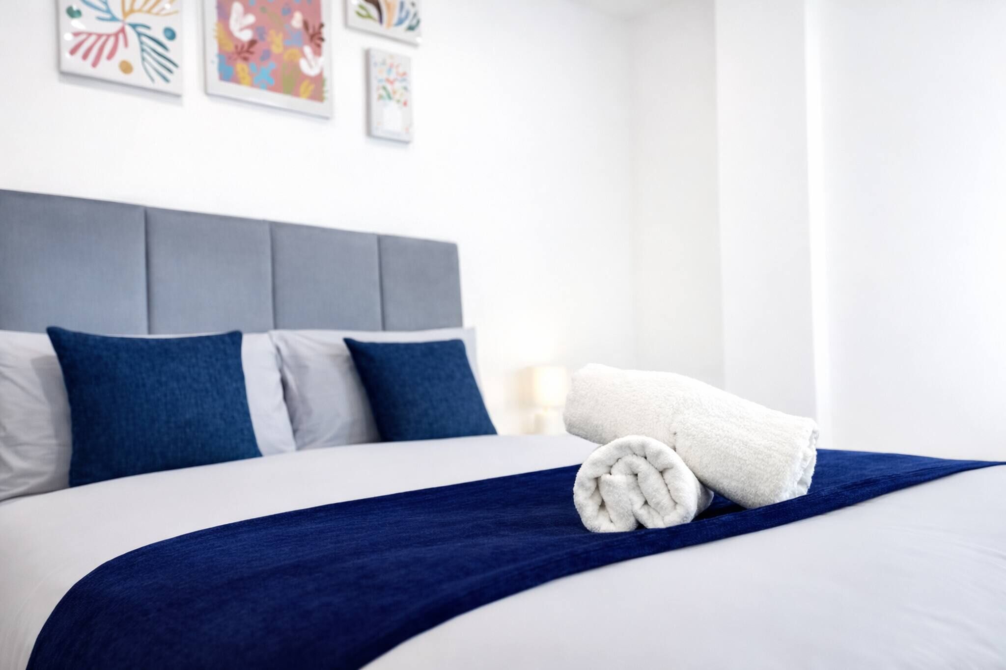 A neatly made bed with a blue blanket and a rolled white towel, a grey headboard, and colorful artwork on the wall.