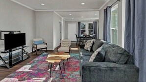 Un salon moderne avec un téléviseur à écran plat, un canapé gris, un tapis à motifs et un coin repas à l’arrière-plan.