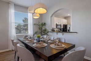 A dining area with a long table set for four, a potted plant, and a kitchen in the background. - WH7712 - 6BR house w/ Resort Access near Disney (Kissimmee)