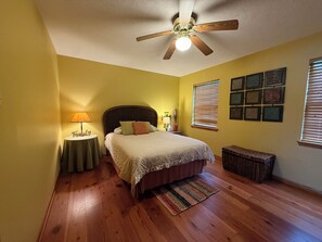 Une chambre à coucher avec un lit, un ventilateur de plafond, une table de chevet avec une lampe, une fenêtre avec des stores et des tableaux encadrés accrochés au mur.