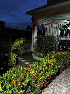 A residential building with a gated entrance, a small garden with red flowers, and a palm tree. - Romantic and tropical apartment in the charming city of Puerto Plata (Puerto Plata)