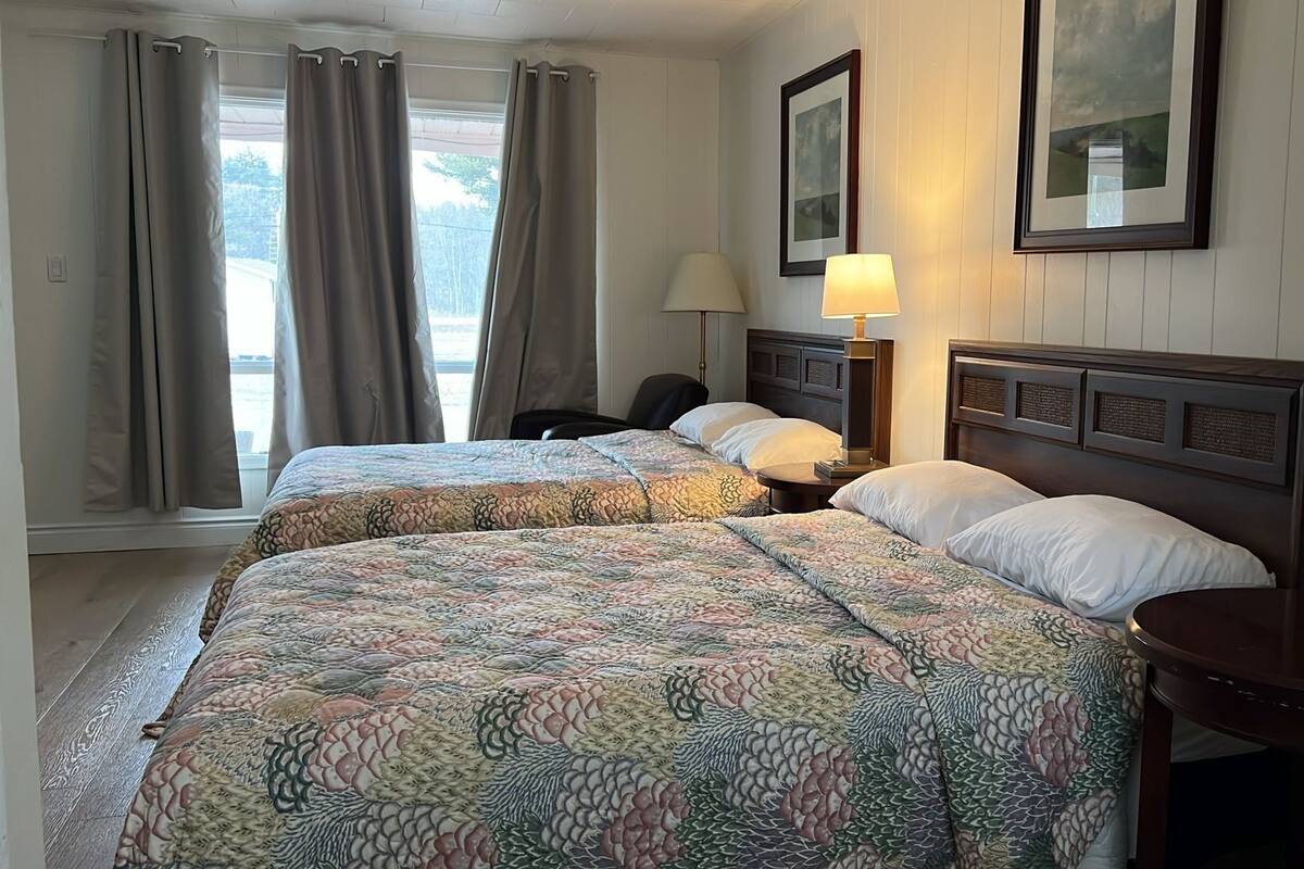 A hotel room with two beds, a nightstand, a lamp, a chair, and a window with curtains.
