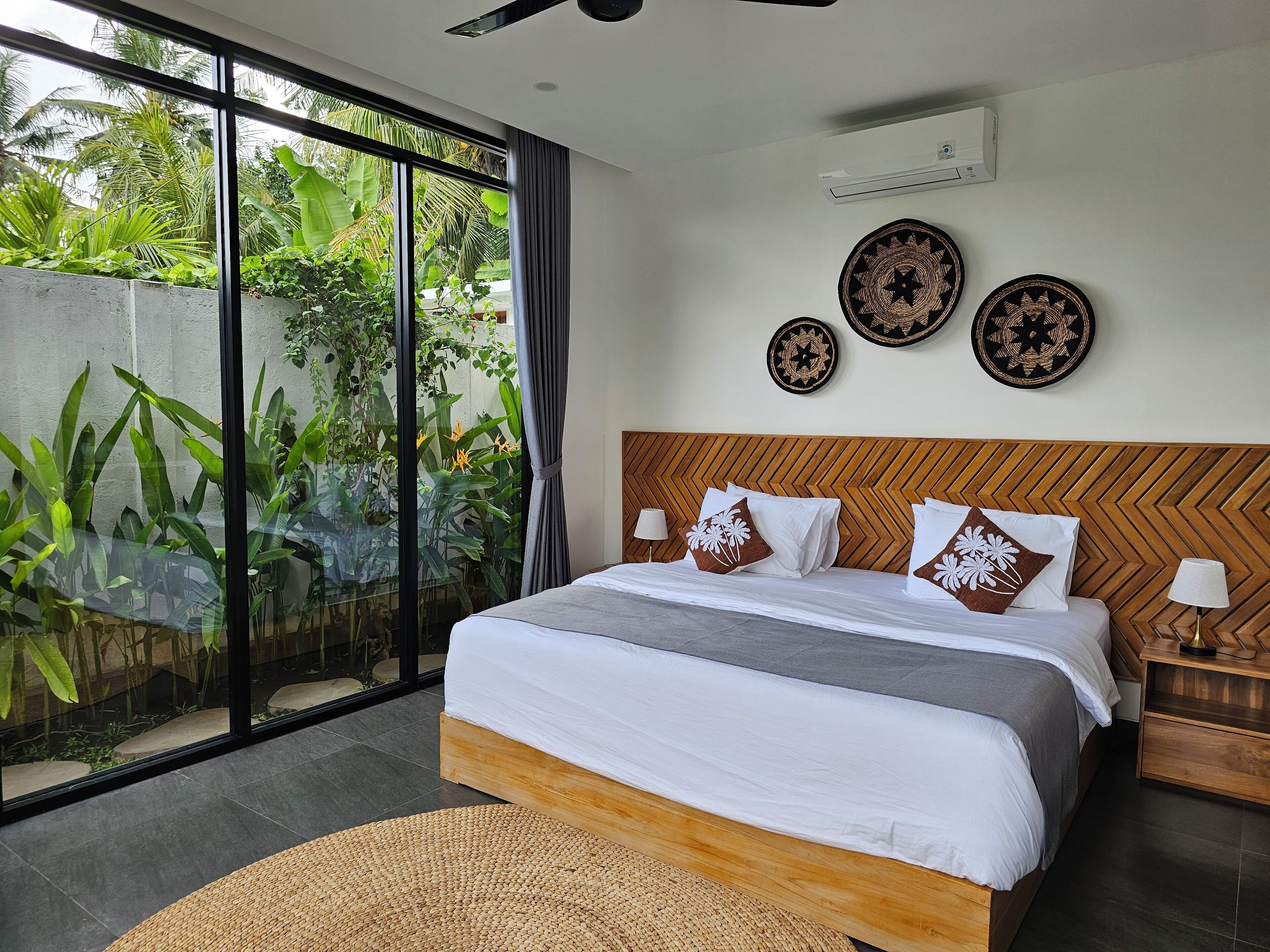 Una camera da letto moderna con un letto grande, vista su piante tropicali attraverso finestre a tutta altezza e pannelli decorativi alle pareti.