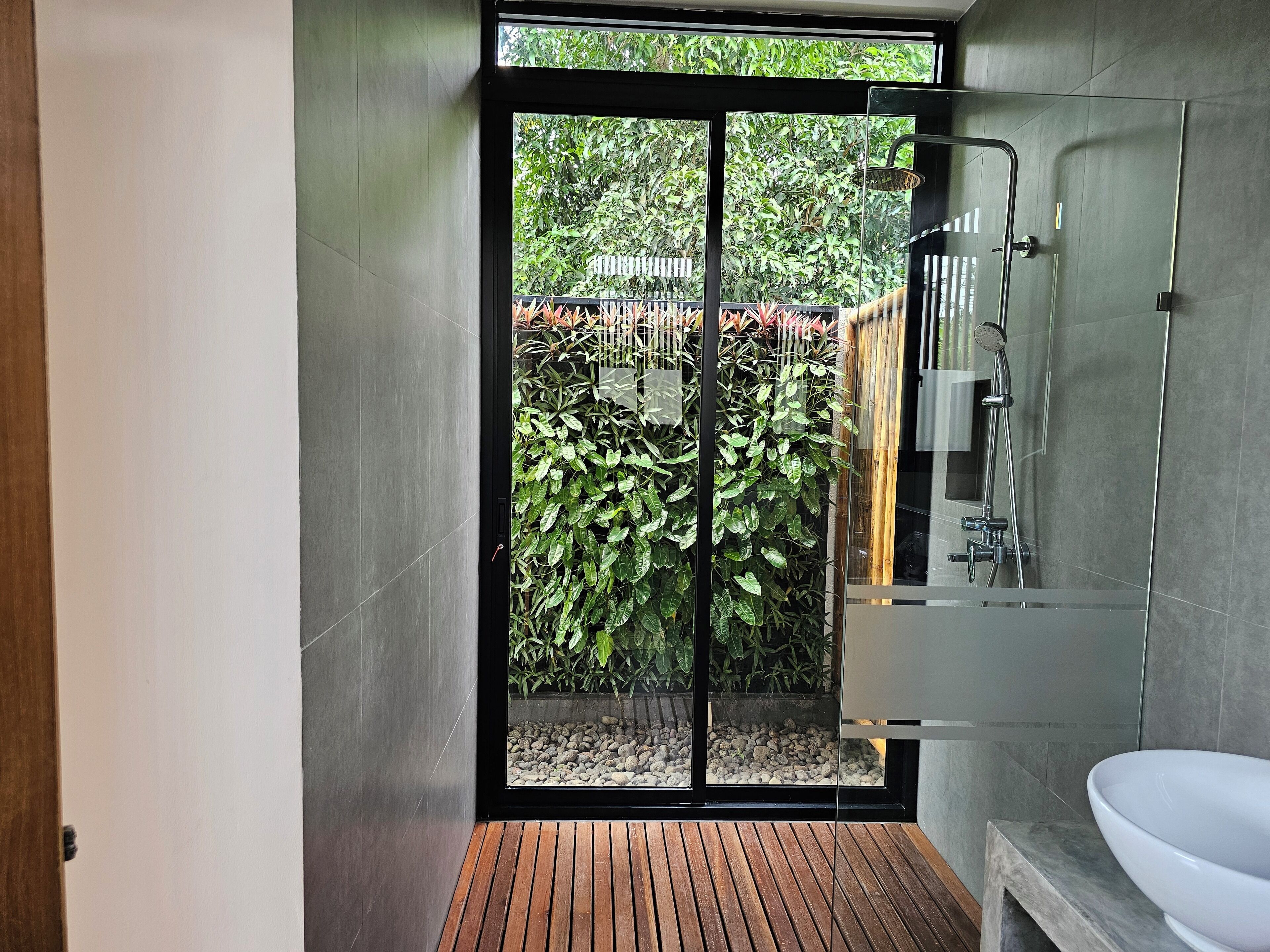 A modern bathroom with a glass shower enclosure, a wooden floor, and a view of a garden through a sliding door.