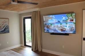 Habitación con una televisión mostrando una escena de playa, una puerta corrediza de vidrio y un ventilador de techo.