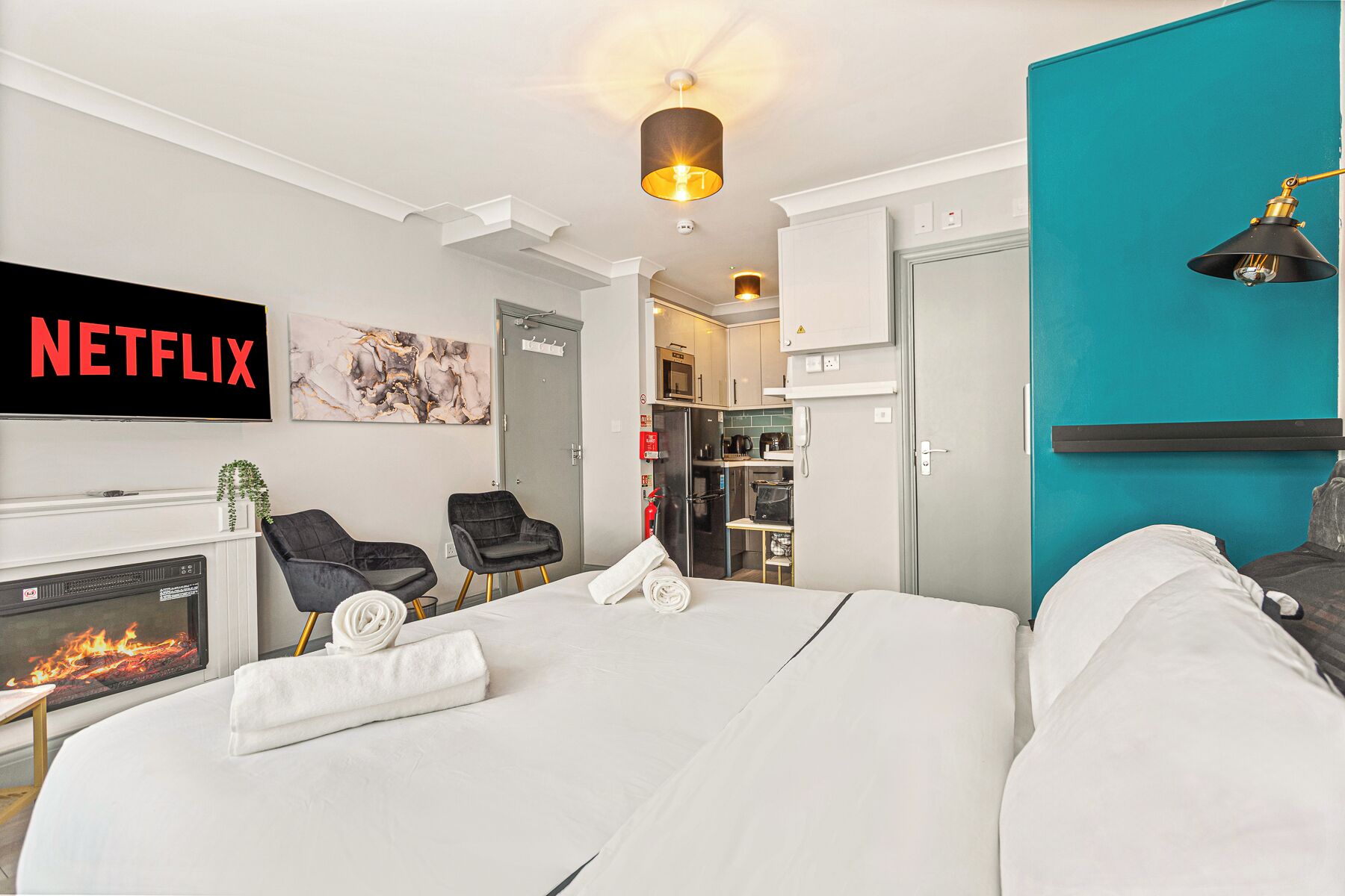 A hotel room with a bed, a TV displaying 'NETFLIX', a fireplace, a kitchen area, and a teal accent wall.