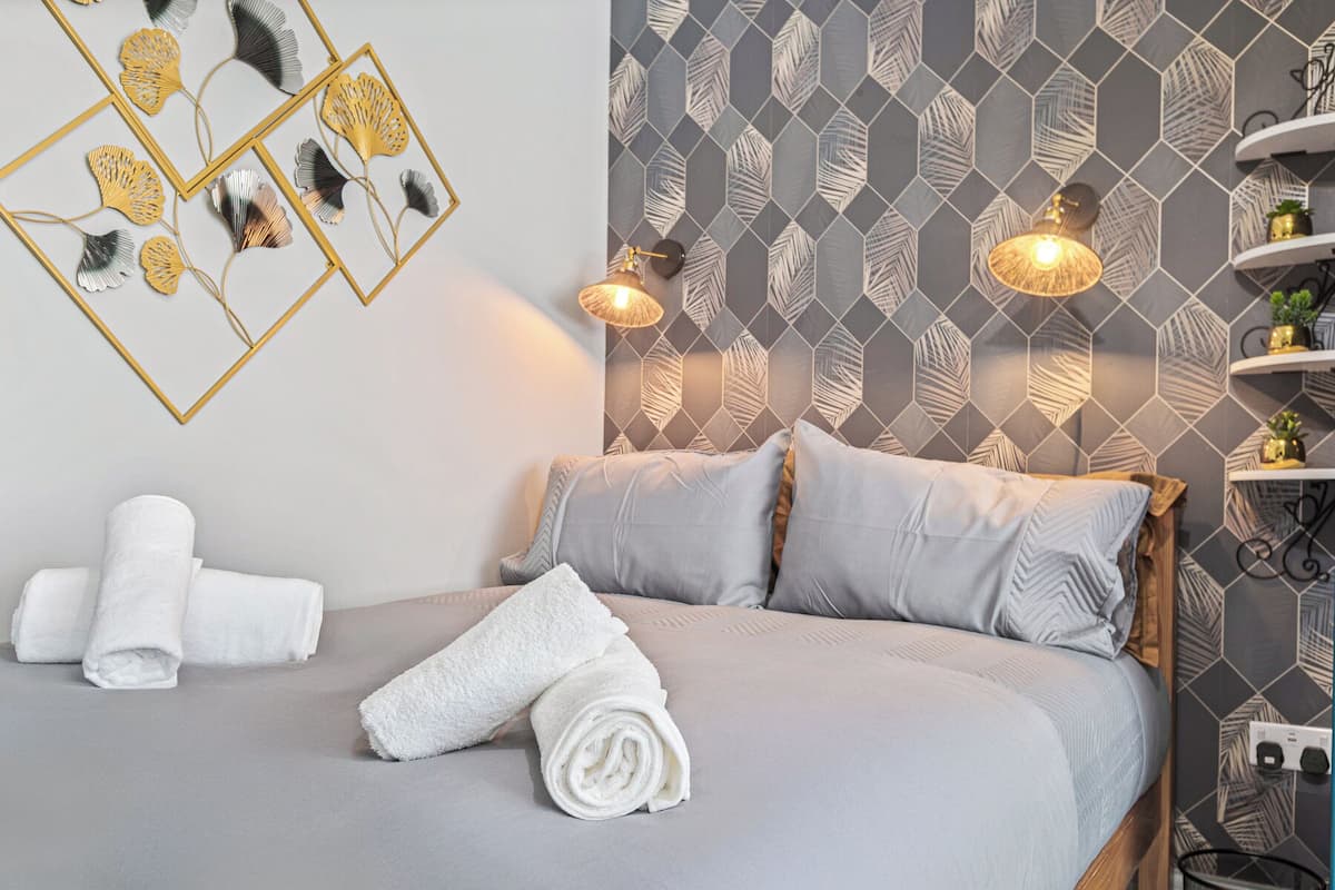 A neatly made bed with pillows and a rolled towel, a wall with geometric leaf-patterned tiles, and two wall-mounted lamps.