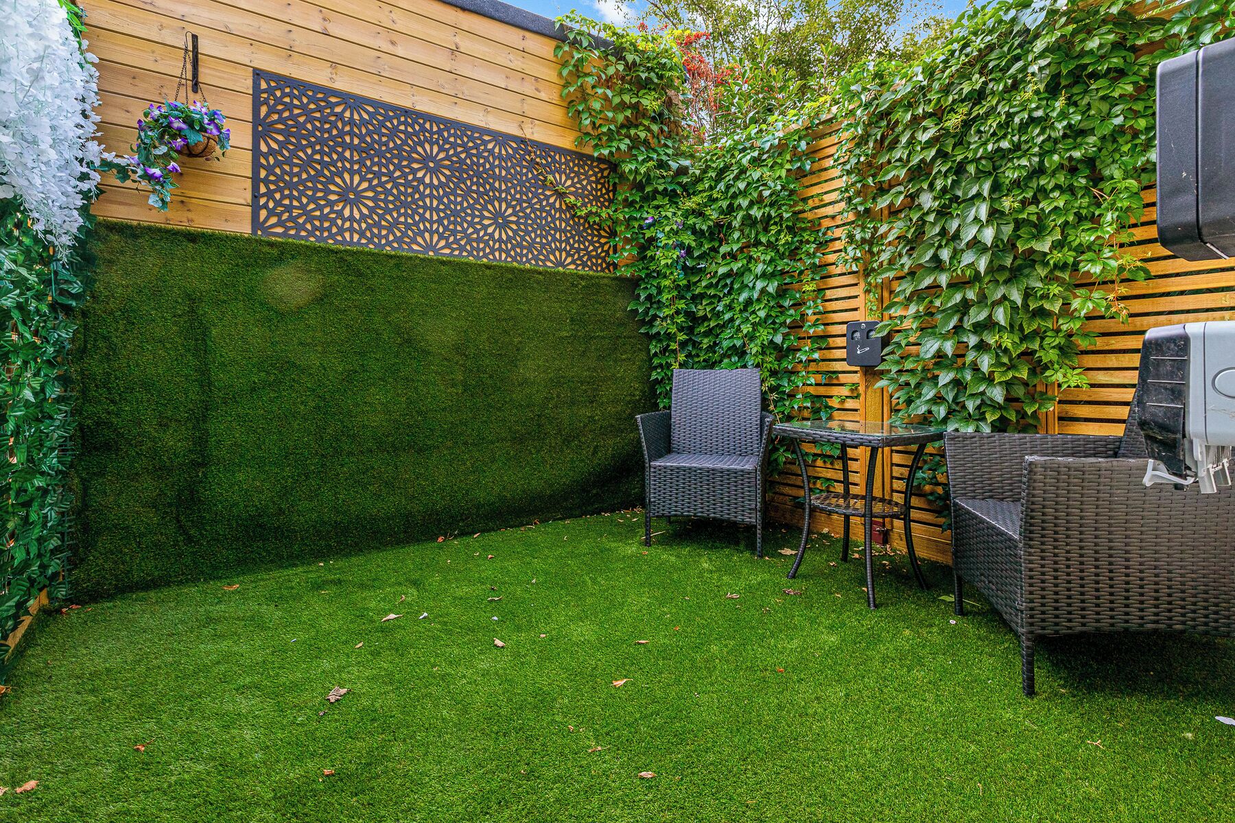 A patio area with a wooden fence, green artificial grass, wicker furniture, and a grill station.