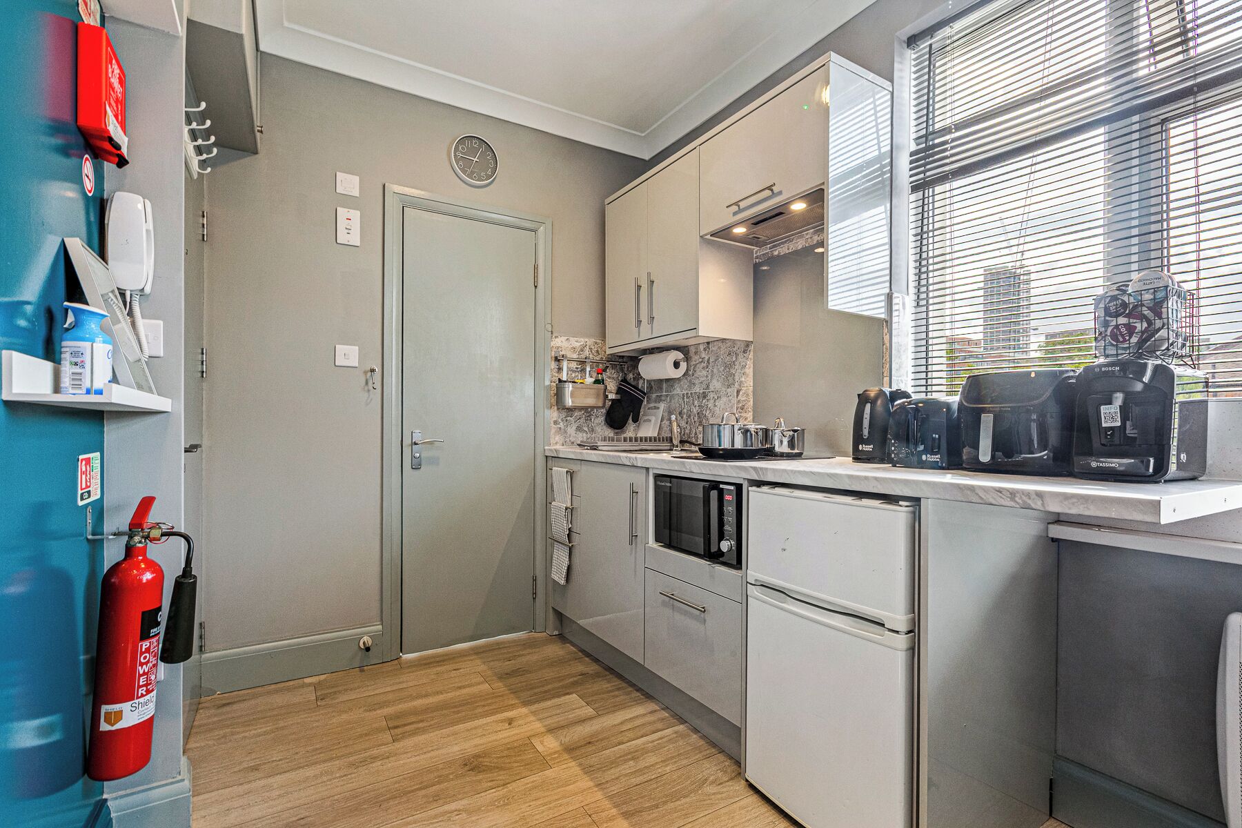 A compact kitchen with a microwave, toaster, and coffee maker on the counter, a fire extinguisher, and a window with blinds.