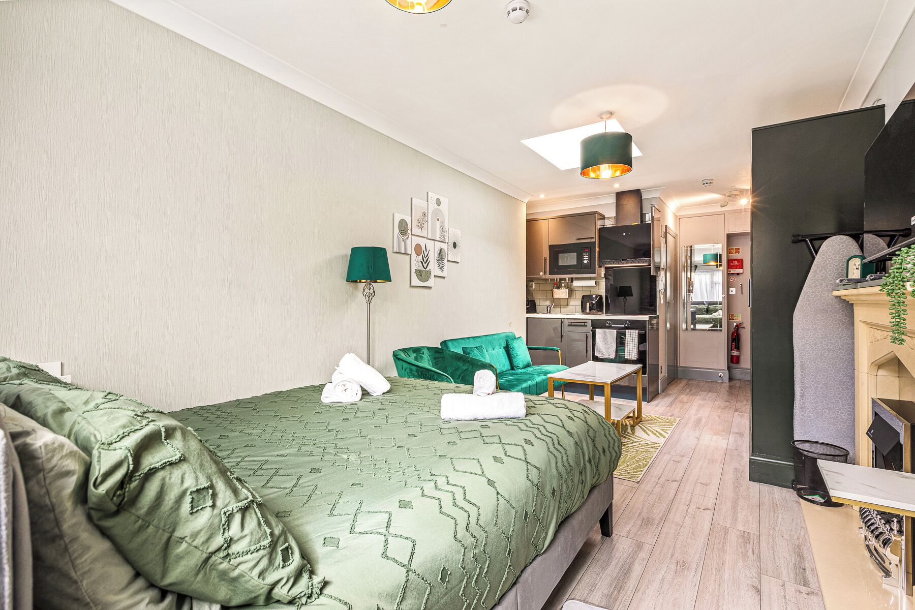 A compact hotel room with a bed, a green armchair, a small table, and a kitchenette in the background.