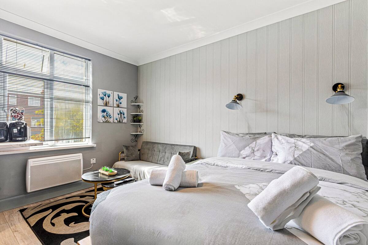 A modern bedroom with a large bed, a bedside table, a radiator, a window with blinds, and a wall-mounted shelf with decorative items.
