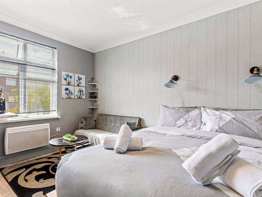 Un dormitorio moderno con una cama grande, una mesita de noche, un radiador, una ventana con persianas y un estante montado en la pared con objetos decorativos.