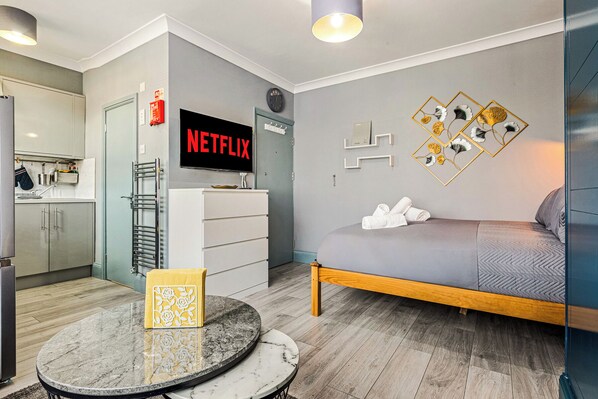 A modern hotel room with a grey sofa, a round marble table, a TV displaying 'NETFLIX', and a wall with geometric floral art. - Imperial Inn Apartments (London)