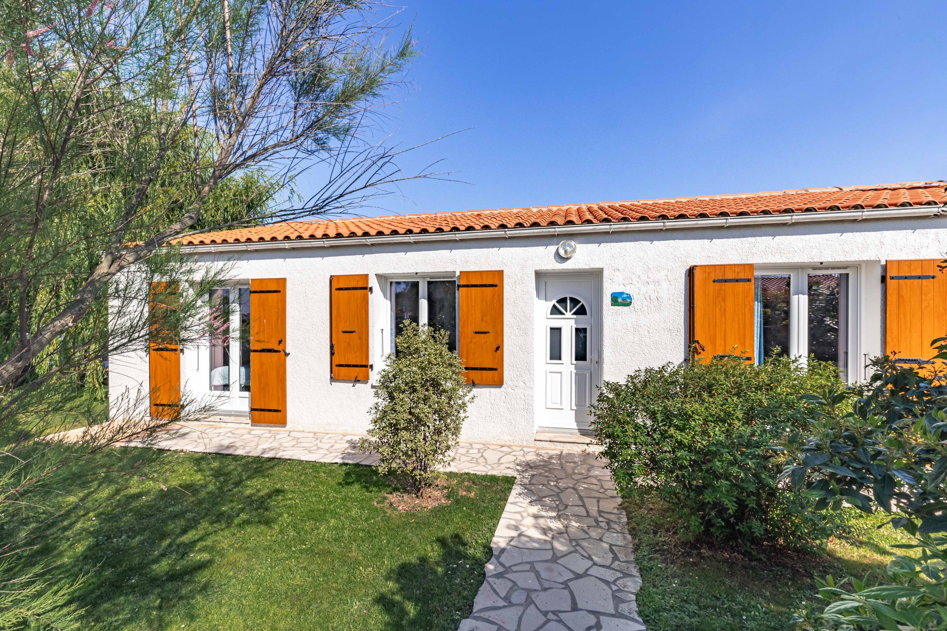Ein weißes Gebäude mit orangenen Fensterläden, einem Steinweg und Grünpflanzen.