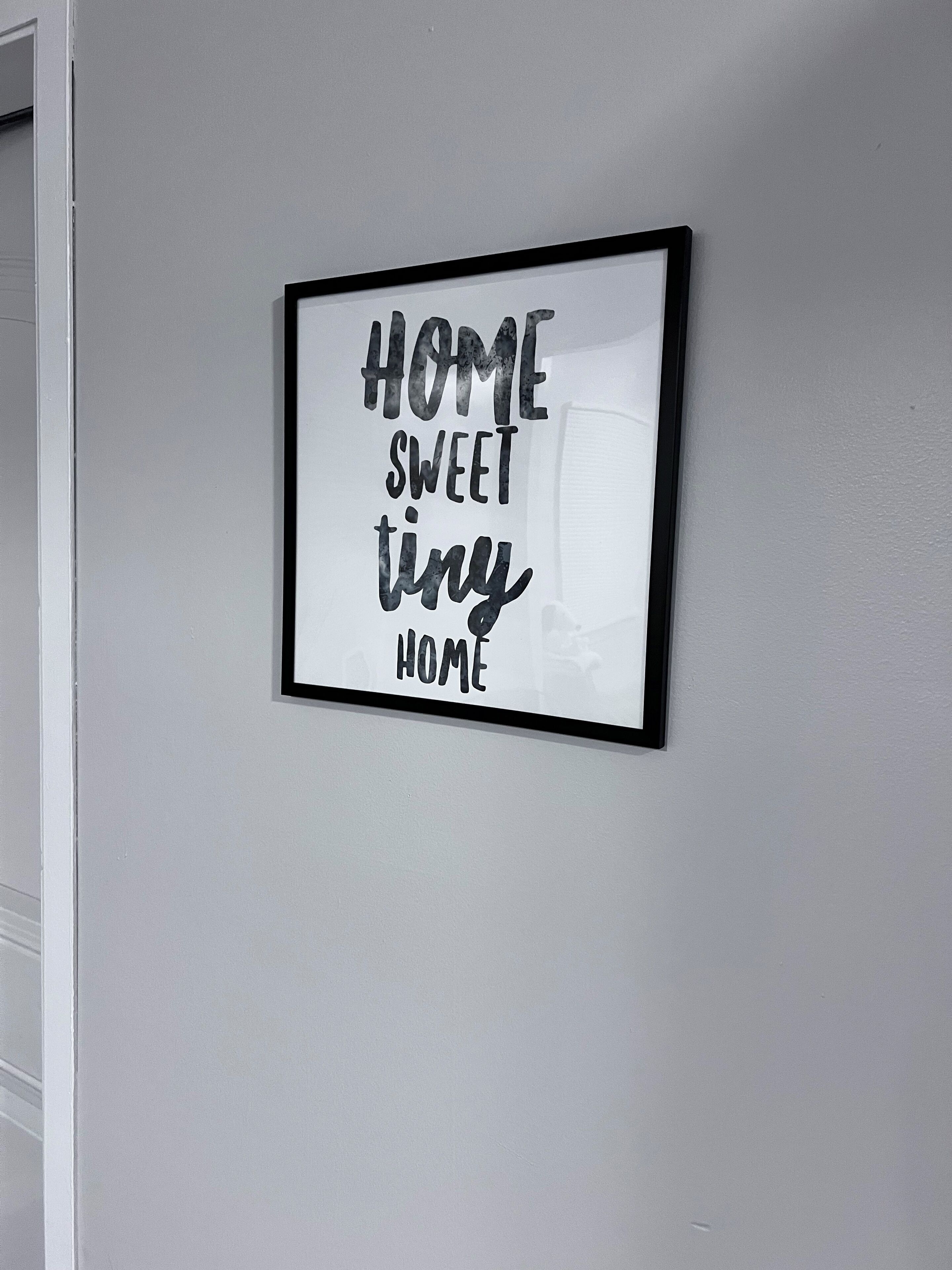 Un cartel enmarcado con el texto 'HOME SWEET TINY HOME' en una pared blanca.