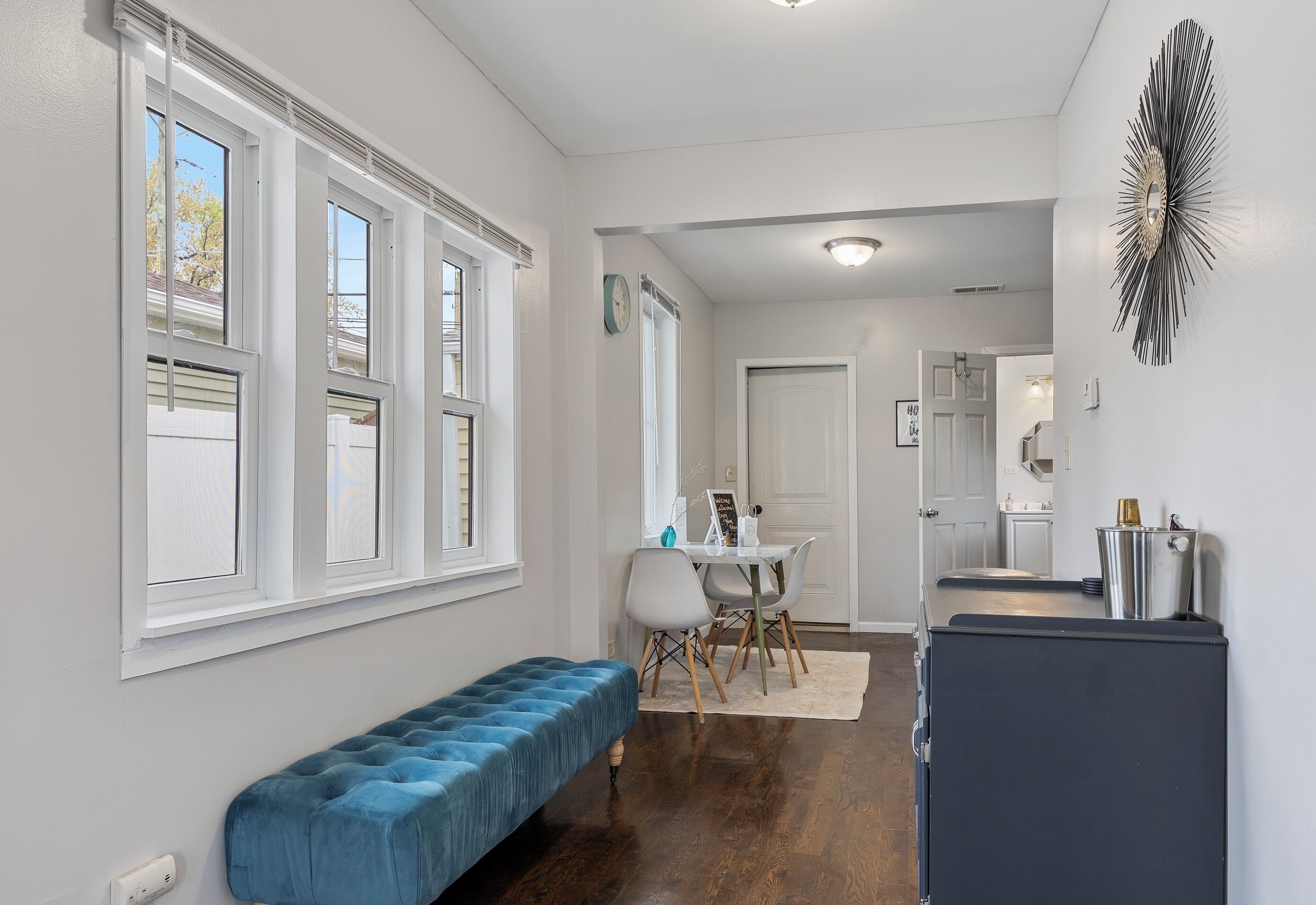 A hallway with a blue bench, a dining area with a white table and chairs, and a kitchen area with a refrigerator.