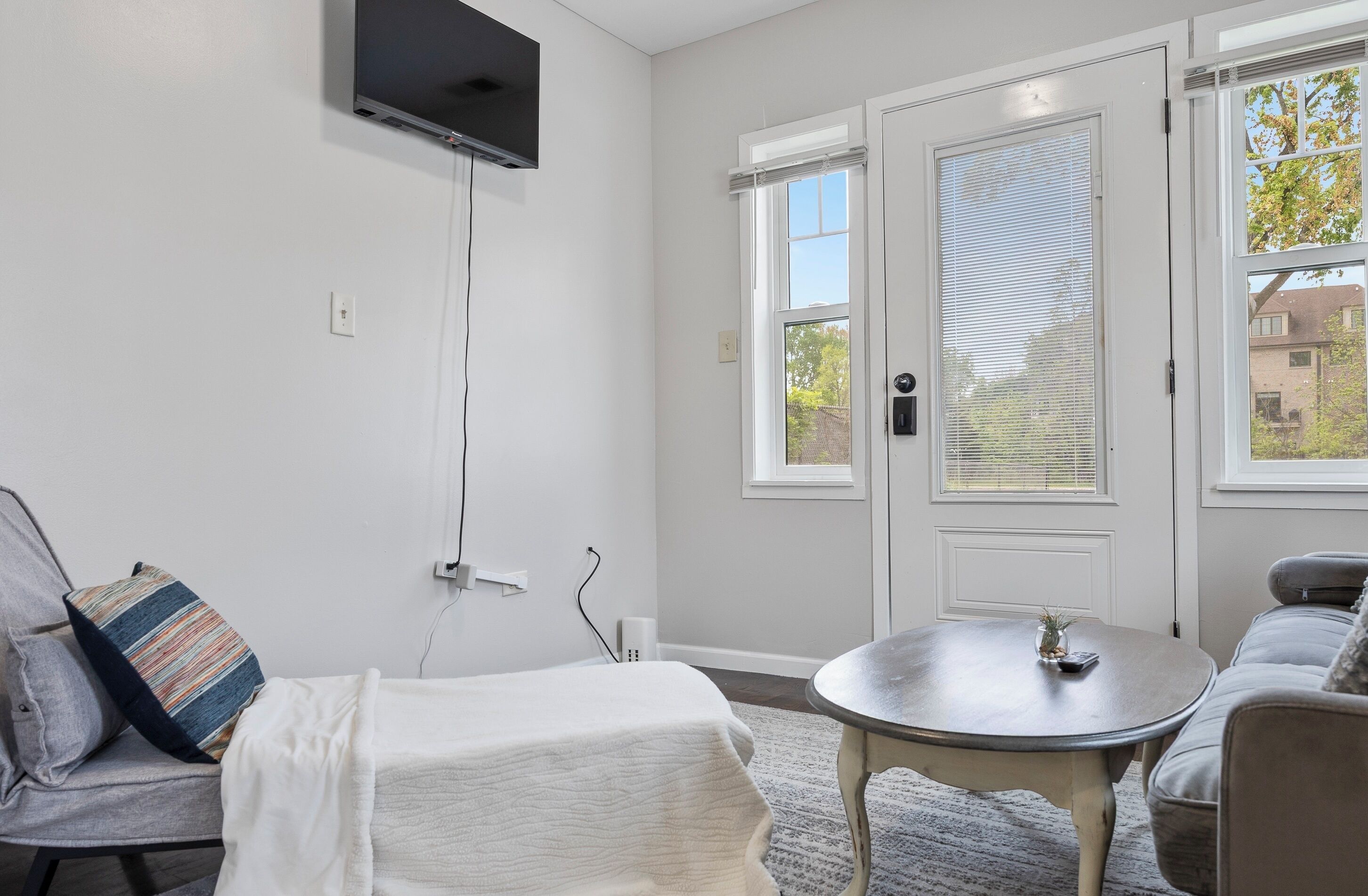 A living room with a sofa, a round table, a TV mounted on the wall, and a door with windows.