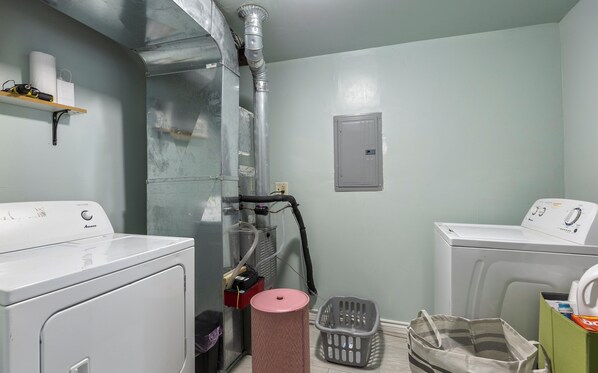 A laundry room with two white washing machines, a metal exhaust vent, a red trash can, a gray laundry basket, and a green storage box.