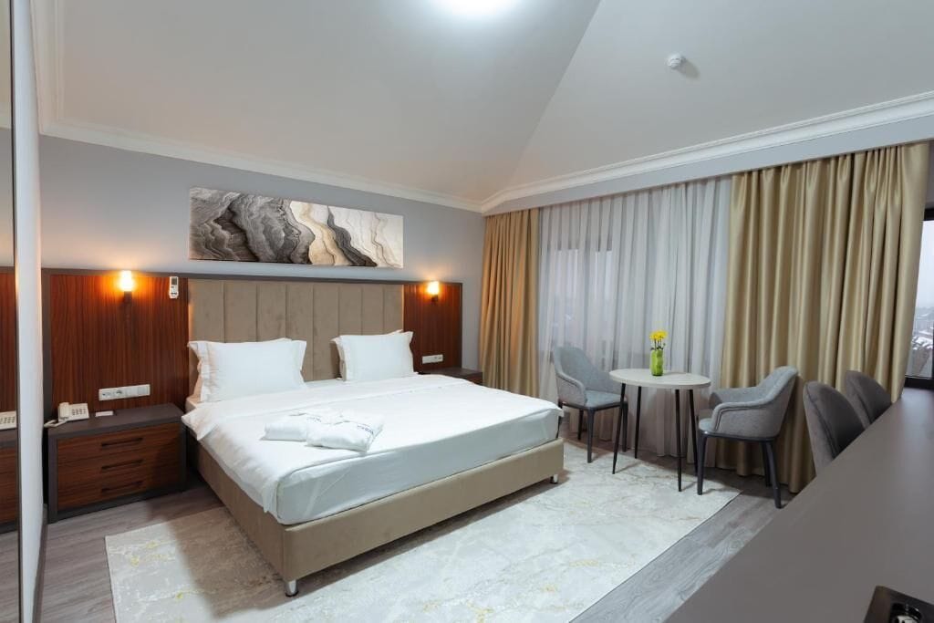 A hotel room with a large bed, bedside tables, a desk with chairs, and a painting on the wall.