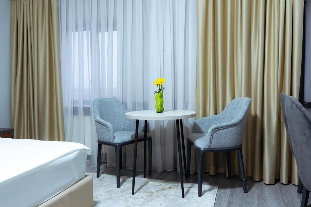 Ett hotellrum med en säng, två grå fåtöljer, ett litet vitt bord med en vas med gula blommor och tunna, genomskinliga gardiner.