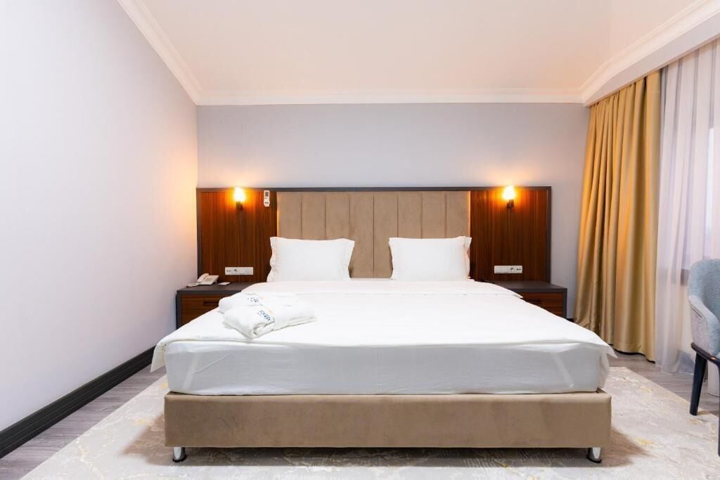 A hotel room with a large bed, two bedside tables, a nightstand, a chair, and a window with curtains.