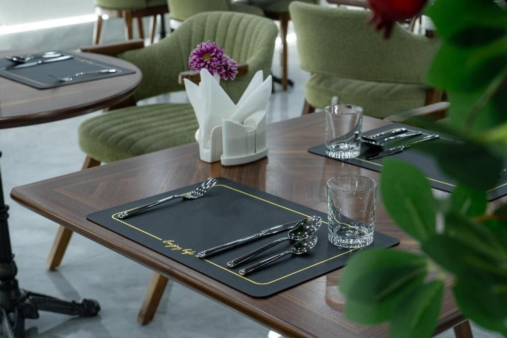 Een eettafel gedekt met een zwarte placemat, bestek, een glas en een witte servethouder met een paarse bloem.