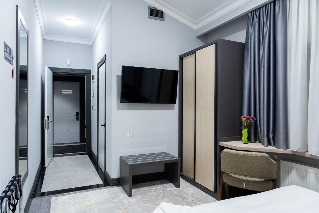 Una habitación de hotel con cama, escritorio con silla, televisor montado en la pared, armario y un pequeño banco.
