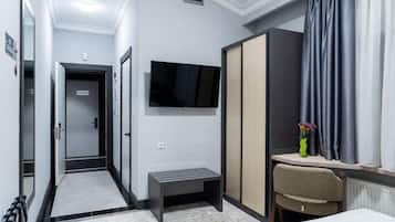 Una habitación de hotel con cama, escritorio con silla, televisor montado en la pared, armario y un pequeño banco.