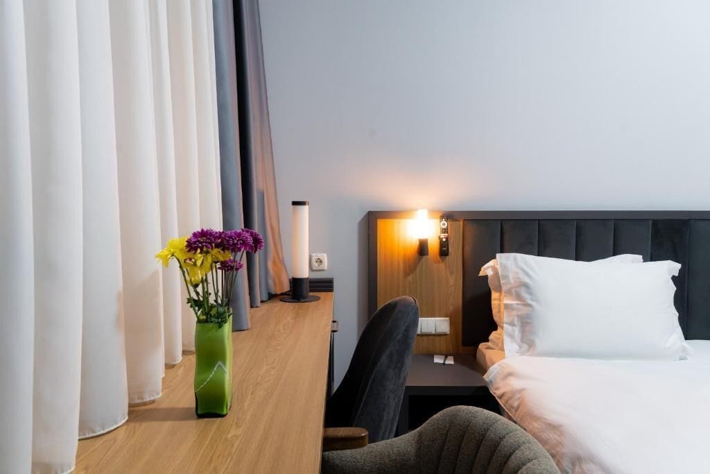 A hotel room with a bed, a desk with a lamp, a chair, and a vase with flowers.