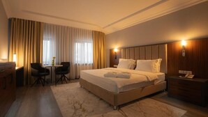 A hotel room with a large bed, a desk with a chair, a small table with a vase, and a nightstand with a lamp. - Rauza Hotel Tashkent (Tashkent)