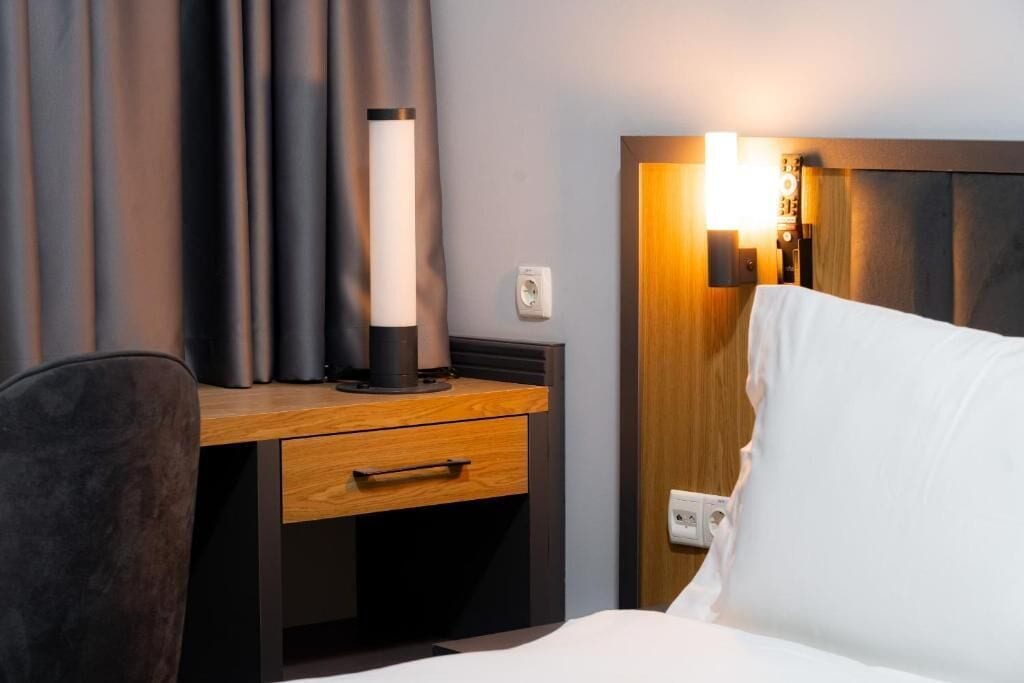 Una camera d'hotel con una scrivania in legno, una lampada, un letto con biancheria bianca e un punto luce a parete.