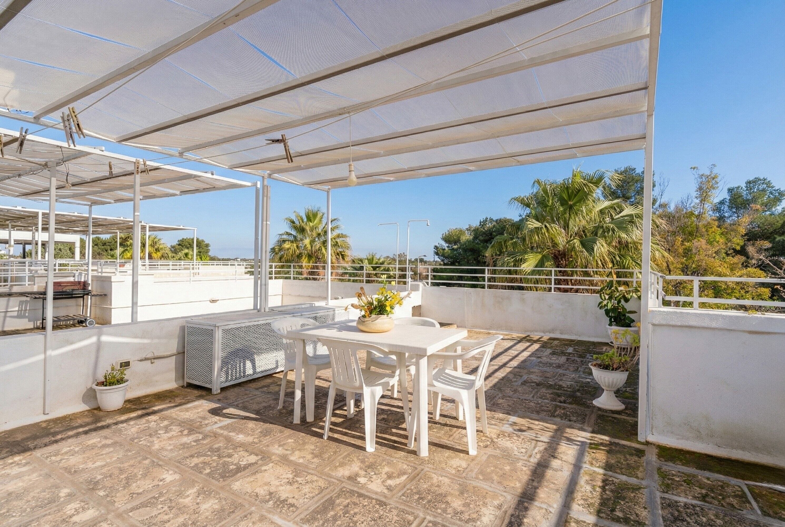 Eine überdachte Terrasse im Freien mit Tisch und Stühlen, umgeben von Blumentöpfen und Blick auf Palmen.