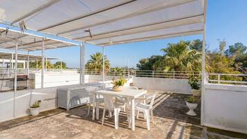 Eine überdachte Terrasse im Freien mit Tisch und Stühlen, umgeben von Blumentöpfen und Blick auf Palmen.