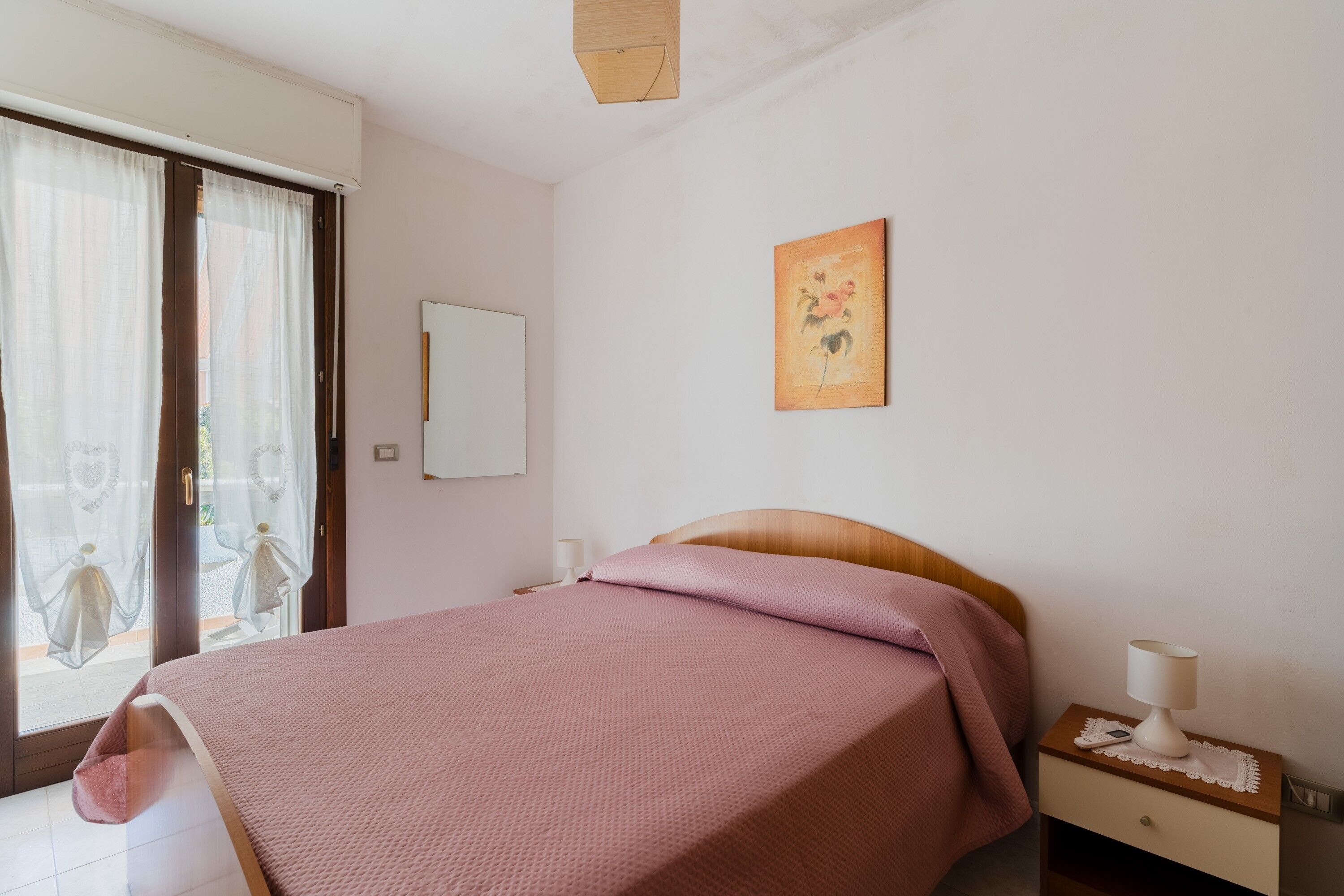 Een slaapkamer met een bed, een nachtkastje met lamp, een raam met gordijnen en een aan de muur hangend schilderij.