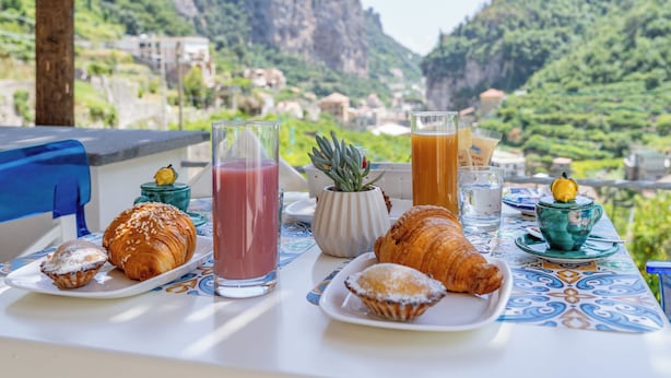 A table with croissants, a glass of juice, and a cup with a lemon on a table with a view of a mountainous landscape. - Villa Zagara (Amalfi)