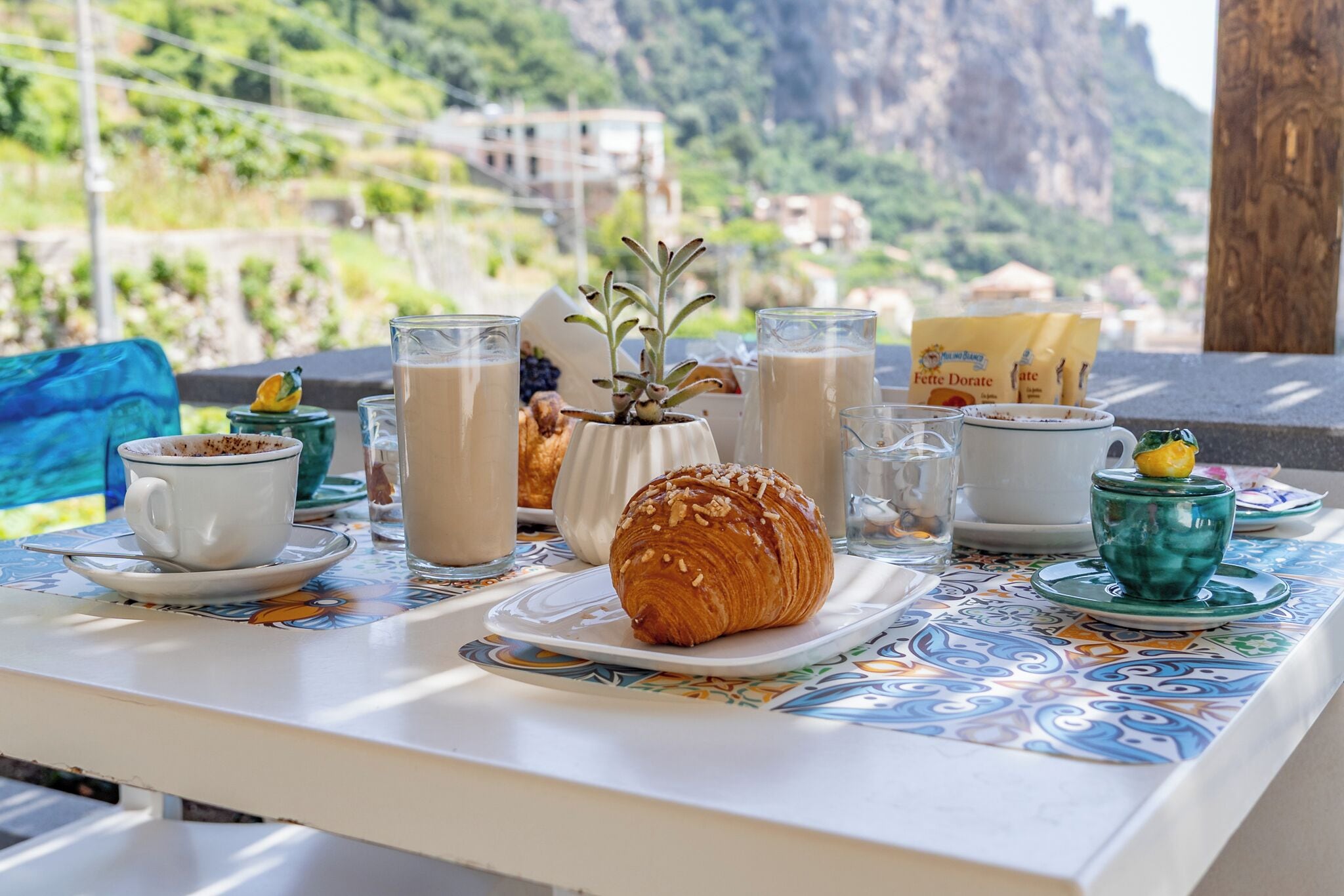 Una mesa con un croissant, tazas de café y una botella de leche, con un paisaje montañoso de fondo.