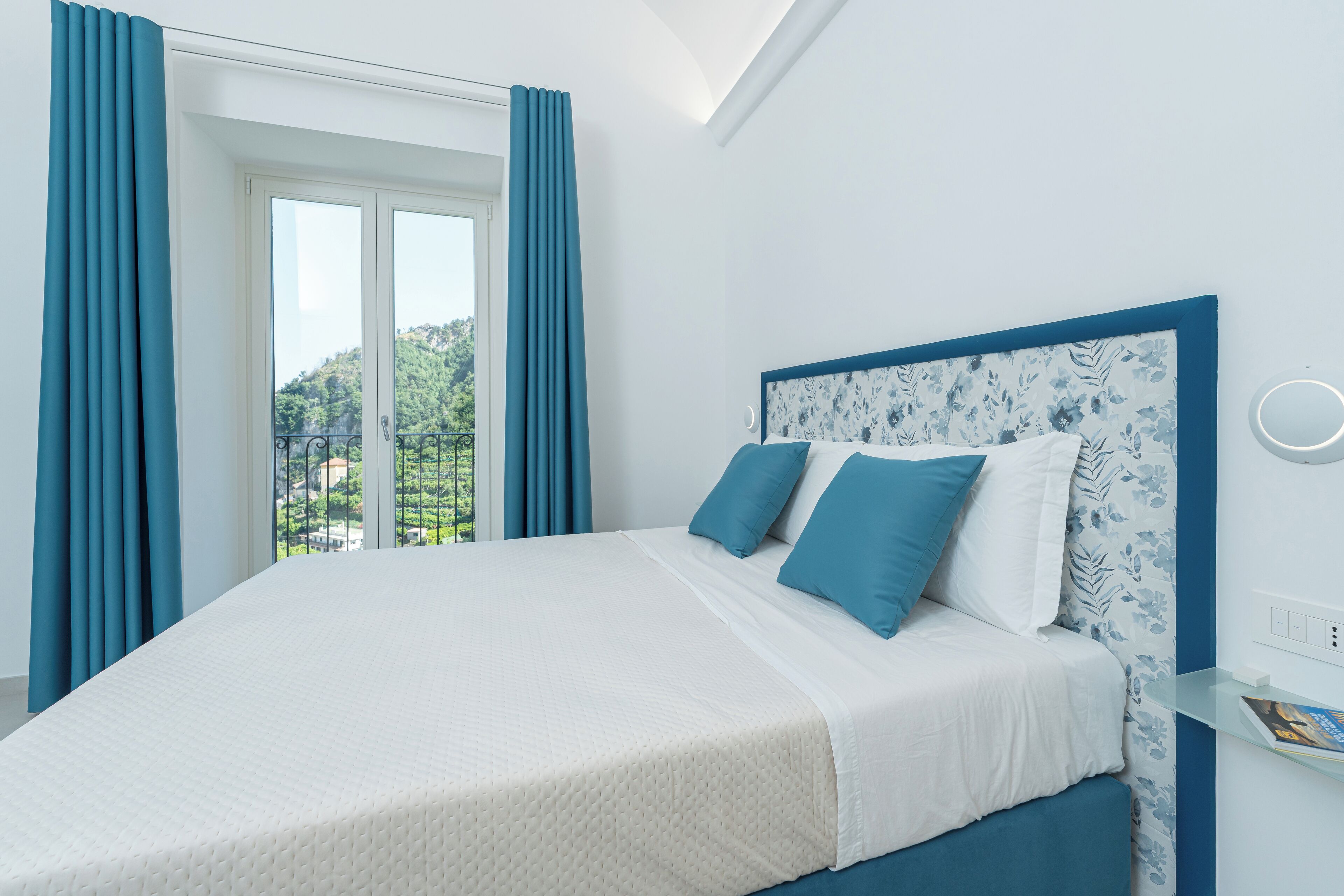 A bedroom with a large bed, blue curtains, and a view of greenery outside.