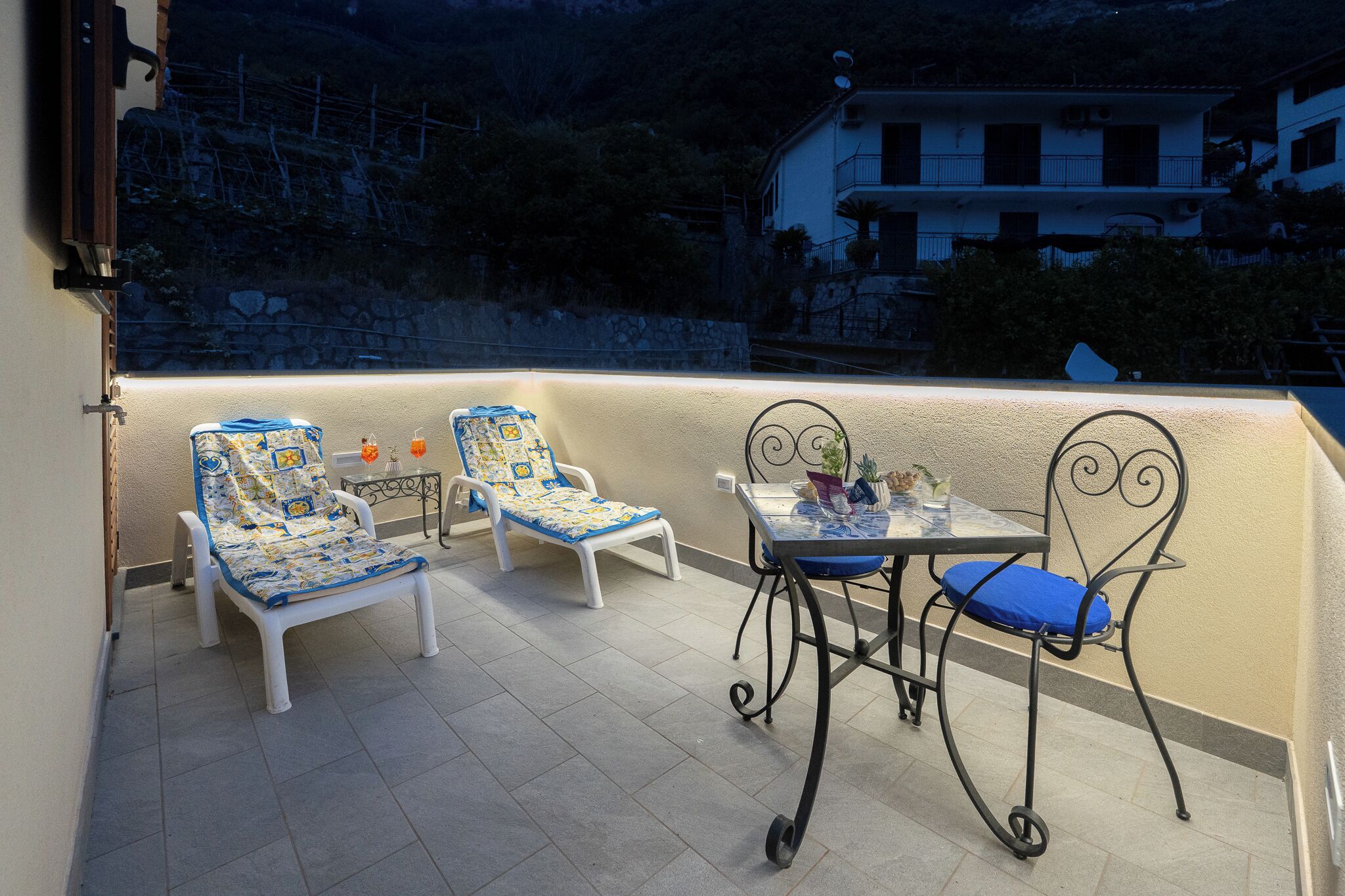 A balcony with two white lounge chairs and a table with chairs, a potted plant, and a small table lamp.