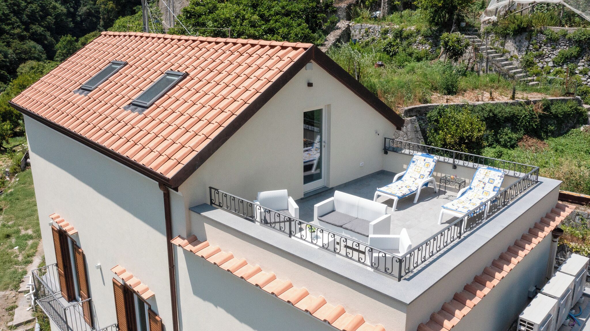 A two-story building with a red-tiled roof, a balcony with white outdoor furniture, and a stone staircase leading to the upper level.