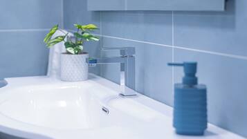 Une salle de bain moderne avec un lavabo blanc, un robinet chromé, un distributeur de savon bleu et une plante en pot.