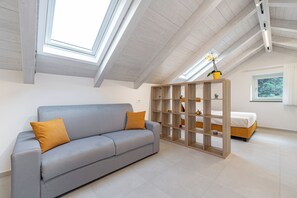 Une chambre de style loft moderne, avec un plafond en pente, une bibliothèque intégrée en bois, un canapé gris agrémenté de coussins orange et un lit avec un matelas blanc.