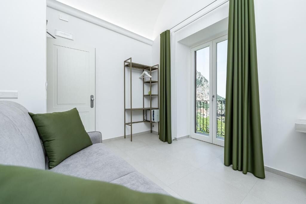 A modern, well-lit room with a grey sofa, a metal shelving unit, and a sliding glass door leading to a balcony.