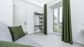 A modern, well-lit room with a grey sofa, a metal shelving unit, and a sliding glass door leading to a balcony.
