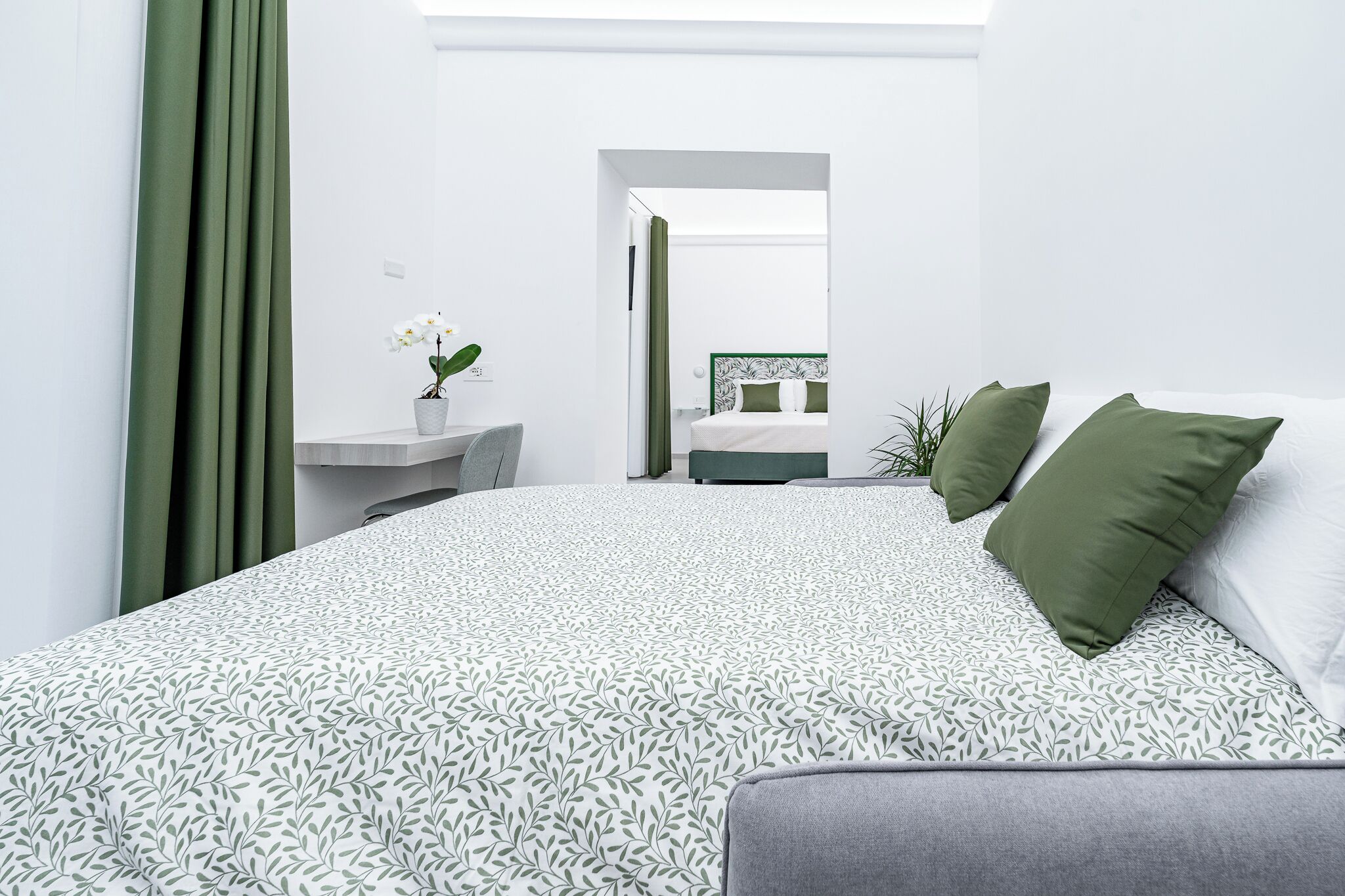 Ein modernes Schlafzimmer mit einem großen Bett, einem Nachttisch mit Pflanze und Blick in einen weiteren Raum.