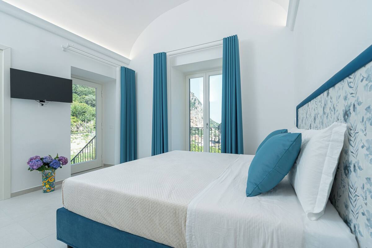 A bedroom with a bed, a TV mounted on the wall, and a balcony with curtains.