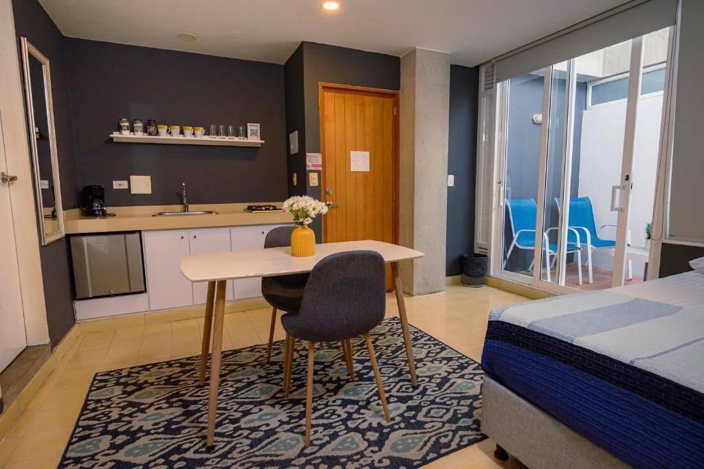 A compact hotel room with a bed, a small kitchenette, a dining area, and a sliding glass door leading to a balcony with blue chairs.